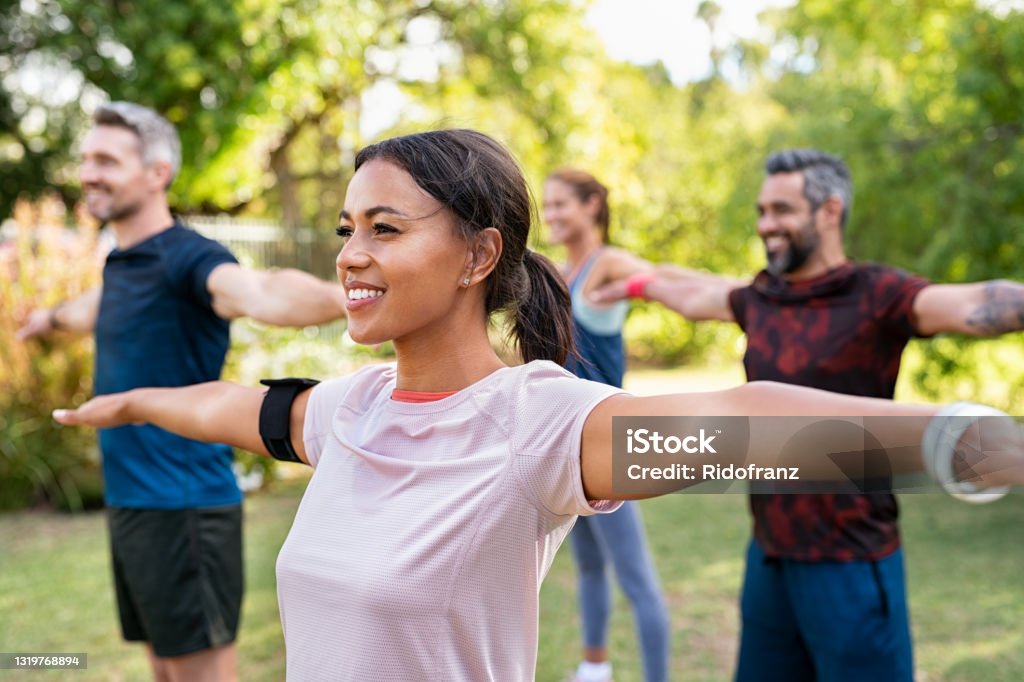 Mixed race woman exercising in park with mature friends Group of multiethnic mature people stretching arms outdoor. Middle aged yoga class doing breathing exercise at park. Beautifil women and fit men doing breath exercise together with outstretched arms. Exercising Stock Photo