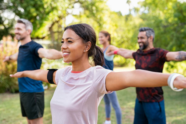 Mixed race woman exercising in park with mature friends Group of multiethnic mature people stretching arms outdoor. Middle aged yoga class doing breathing exercise at park. Beautifil women and fit men doing breath exercise together with outstretched arms. cooling down stock pictures, royalty-free photos & images