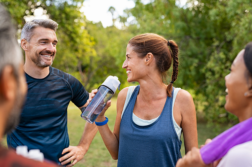 Sporty middle aged woman drinking water from bottle after fitness exercise. Mature group of multiethnic friends have a conversation after fitness training at park. Latin woman drinking water for refreshment after workout in park while talking and smiling with a group of people.