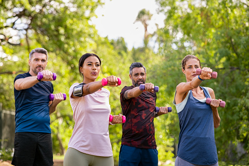 Group of mature friends exercise using dumbbells at park. Team of four middle aged people lifting weights outdoor. Determined men and fit women using dumbbell for workout in park together.