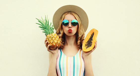 Summer portrait of young woman with pineapple and papaya wearing straw hat, sunglasses on a white background