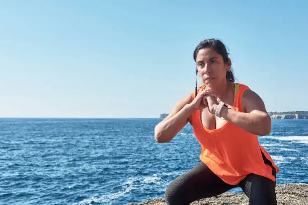 Latin woman, middle-aged, wearing sportswear, training, doing physical exercises, plank, sit-ups, climber's step, burning calories, keeping fit, outdoors by the sea, wearing headphones, smart watch