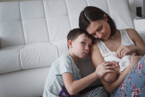 A young mother gently cuddles her newborn in the home interior. Her eldest son is sitting next to her, stroking the baby's head.