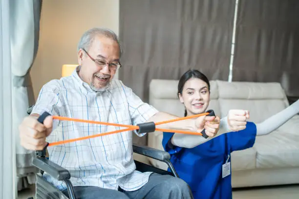 Photo of Asian Disabled senior elderly man on wheelchair doing physiotherapist with support from therapist nurse. Older handicapped man using resistance stretch band exercise for patient in home nursing care