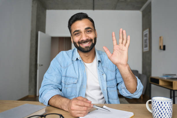 Young happy indian teacher waving hand having video call at home. Webcam view. Young happy indian Hispanic arab teacher talking on video conference call greeting waving hand using laptop at modern home office. Remote distant online work education techs concept. Webcam view. webcam view man stock pictures, royalty-free photos & images