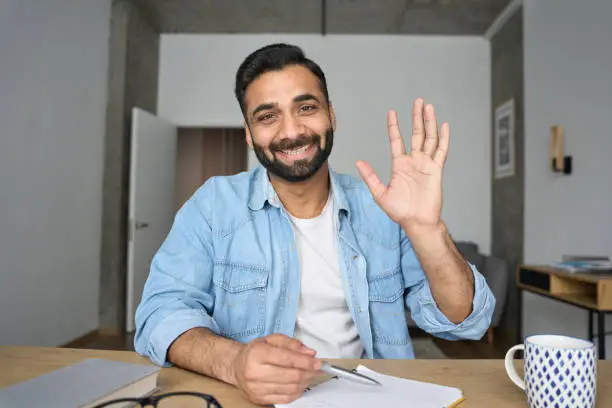 Photo of Young happy indian teacher waving hand having video call at home. Webcam view.