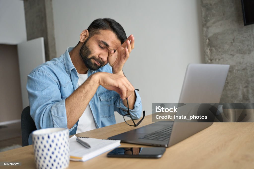 Young tired Hispanic indian student rubbing eyes sitting at desk with laptop. Young indian eastern tired exhausted business man rubbing eyes sitting in modern home office with laptop on desk. Overworked burnout academic Hispanic student with glasses in hand feeling eyestrain. Tired Stock Photo
