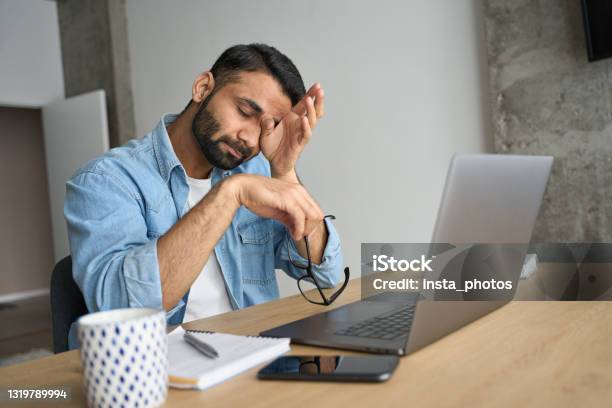 Young Tired Hispanic Indian Student Rubbing Eyes Sitting At Desk With Laptop Stock Photo - Download Image Now