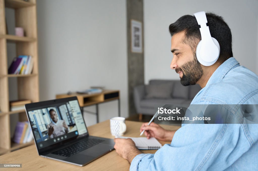 Indian latin male student having video call with male teacher on laptop. Latin indian adult student wearing headset having virtual meeting online call training educational webinar chatting with teacher at home office writing notes. Video e learning conference call on pc. Video Call Stock Photo