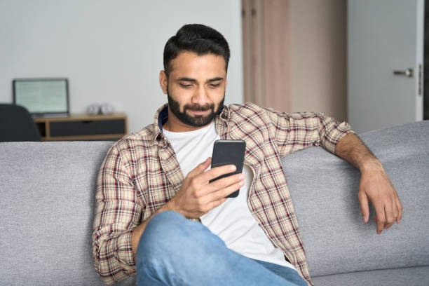 istock Young attractive indian man sitting on sofa holding cellphone at home. 1319790032