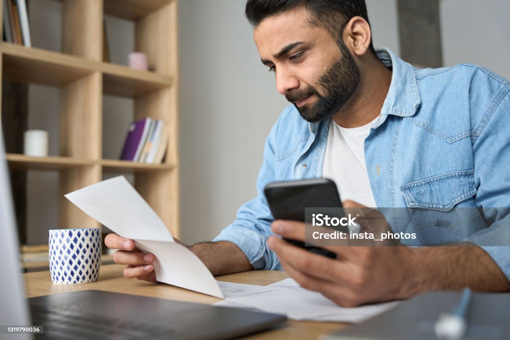 Young indian business man holding phone reading bank receipt calculating taxes. Young indian businessman holding phone reading bank receipt calculating taxes, ethnic man using smartphone mobile application checking bill document, managing money finances, loan expenses. Financial Bill Stock Photo