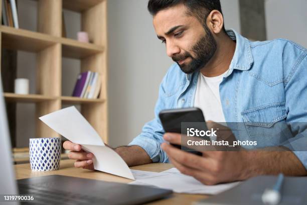 Young Indian Business Man Holding Phone Reading Bank Receipt Calculating Taxes Stock Photo - Download Image Now