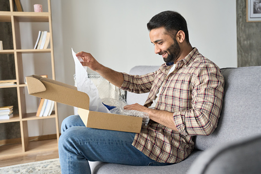 Happy smiling latin indian man opening box with ordered goods at home on couch. Online shopper male customer opening online shop parcel. International delivery service comfort concept.