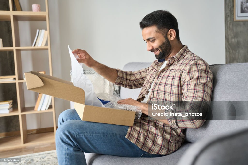 Young happy adult indian man opening parcel box at home on the couch. Happy smiling latin indian man opening box with ordered goods at home on couch. Online shopper male customer opening online shop parcel. International delivery service comfort concept. Box - Container Stock Photo