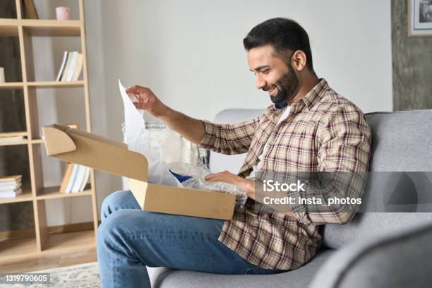 Young Happy Adult Indian Man Opening Parcel Box At Home On The Couch Stock Photo - Download Image Now