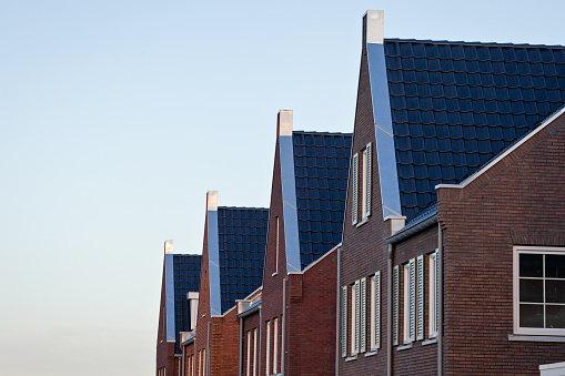 Side view close-up of rooftops of newly built modern detached Dutch brick houses with gable roofs, built with red bricks, window shutters and black roof tiles
