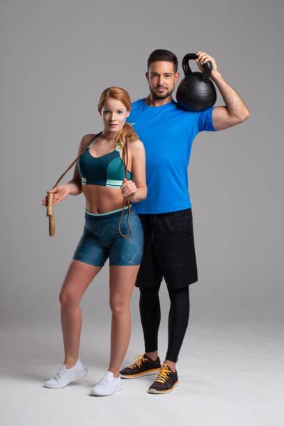 Fit Caucasian couple posing with jump rope and kettlebell in studio on gray background