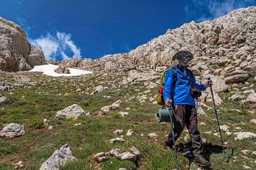 Model in her 60s with backpack and walking pole walking in high position in mountainous area. Wearing sunglasses. Fully equipped hiker. She has a hat on her head.