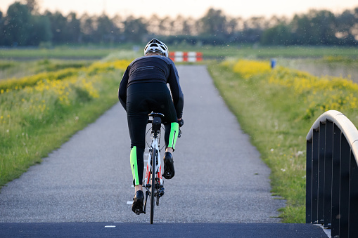 Zevenhuizen, Zuidplas, South Holland, Netherlands, may 18th 2021, rear view of a male cyclist on a road racing bicycle, speeding on an asphalt road in a rural area at sunset after just having crossed a bridge - the air is filled with flies, attracted by the water