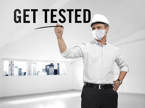 Architect wearing a medical mask writing “Get tested” on air in an empty building