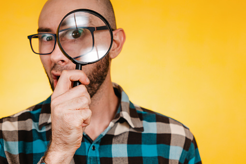 The concept of finding and solving problems. A bald man with a beard and glasses, wearing a blue checked shirt, looks through a magnifying glass. Eye in increased size. Yellow background. Close up and copy space.