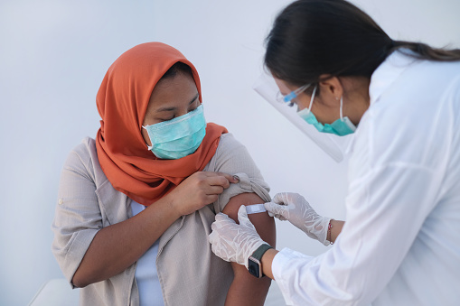 Close-up shot of Asian female doctor putting adhesive bandage to patient's arm. The doctor wearing a face shield, surgical mask and protective gloves for safety protection while the patient are seating on a chair , wearing an orange hijab.