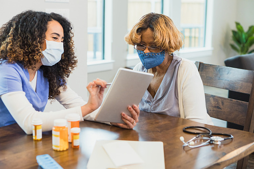 A female nurse shows a female patient a medical document on a digital tablet during a home healthcare visit. The nurse and patient are wearing protective face masks during the COVID-19 pandemic.