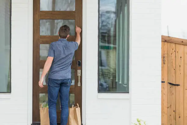 A delivery person knocks on the door of a customer's home. He is leaving the contactless delivery order on the customer's doorstep.