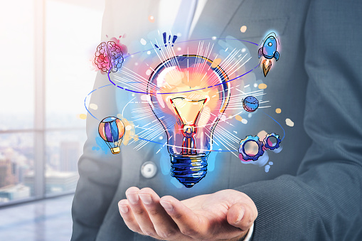 Businessman hand holding hologram of colorful light bulb as a concept of new idea for start up. Concept of creativity and brainstorming. Panoramic office on background