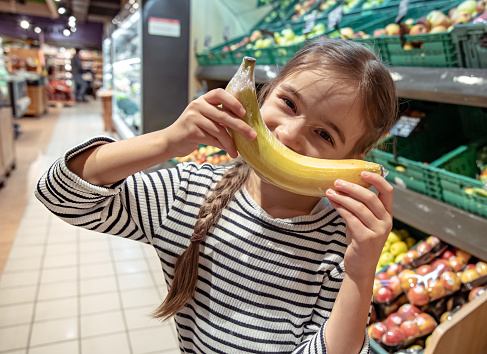 Funny little girl with banana at the supermarket. Individual packaging of fruits.