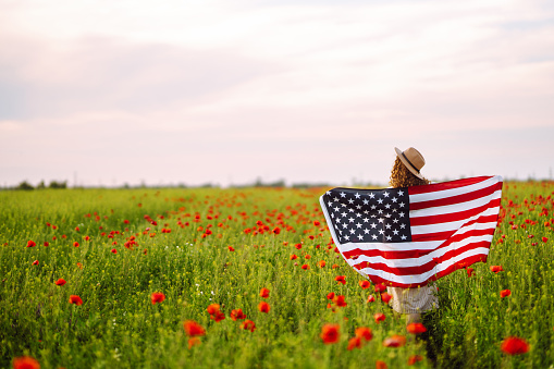 Young woman proudly hold waving american USA flag in in the poppy field. Patriot raise national american flag against the blue sky. Independence Day, 4th July.