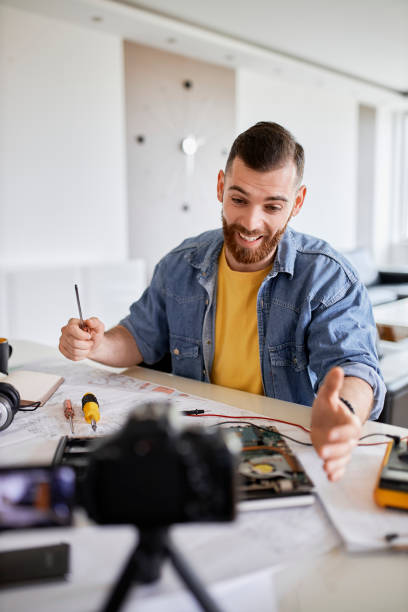 istock Male influencer in workshop doing his day job and filming tutorial. Working table full of different tools and items needed for repairing electrical circuits 1319841371