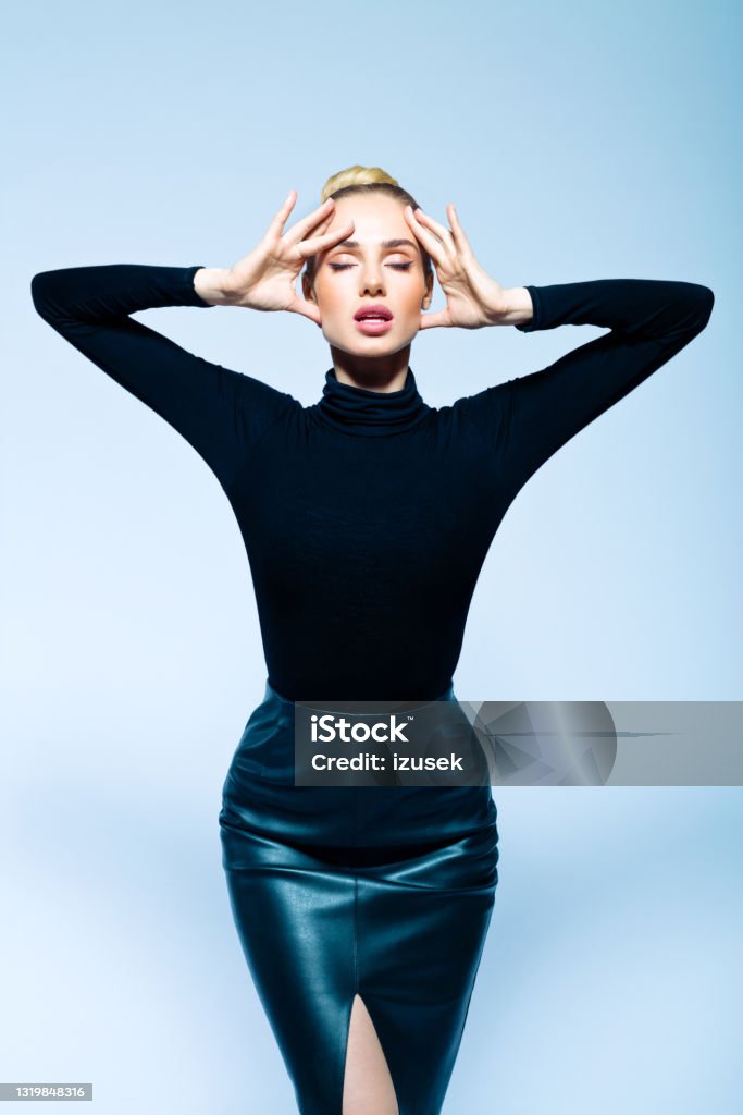 Fashion portrait of confident elegant woman Confident woman wearing black turtleneck and leather skirt standing with raised arms and eyes closed. Studio shot on blue background. Fashion Model Stock Photo