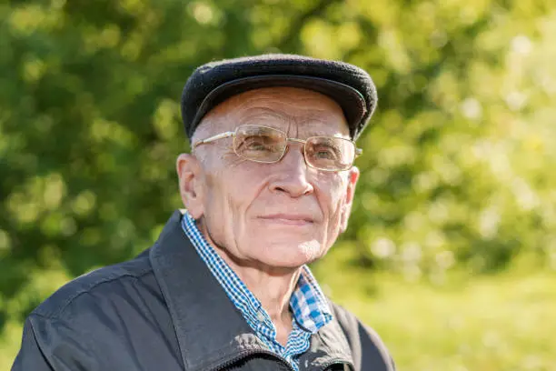 Large scale head portrait of senior man wearing cap and glasses outside on green natural background