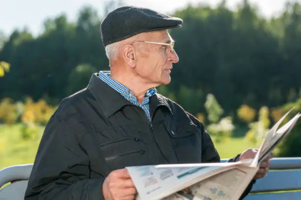 Elder man sitting on wooden bench and wearing cap and glasses holds newspaper in hands outdoor
