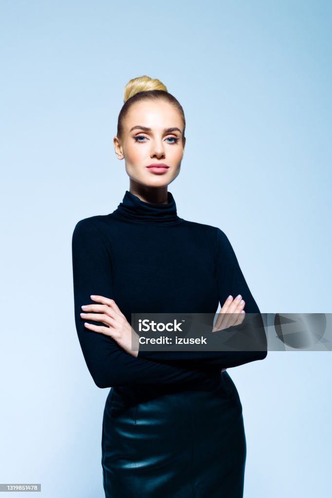 Portrait of confident elegant woman Confident woman wearing black turtleneck and skirt standing with arms crossed and looking at camera. Studio shot on blue background. One Woman Only Stock Photo