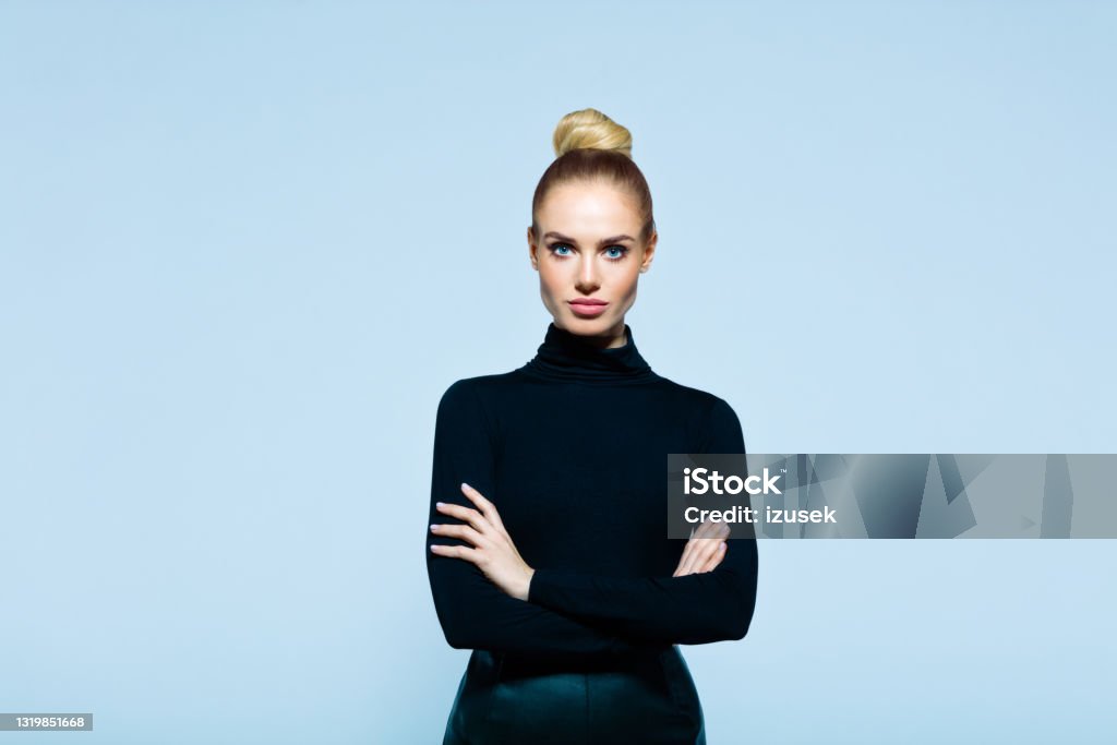 Portrait of confident elegant woman Confident woman wearing black turtleneck and skirt standing with arms crossed and looking at camera. Studio shot on blue background. Females Stock Photo
