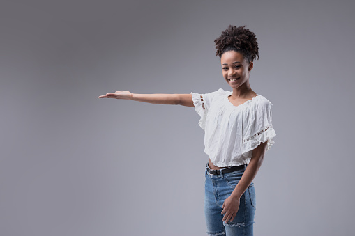 Attractive young Black girl in jeans holding out her empty palm for product placement for advertising with a happy friendly confident smile on a grey studio background with copyspace