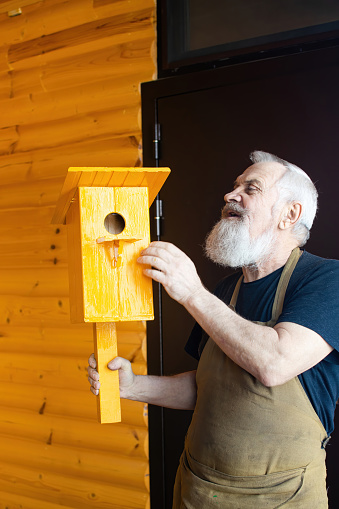 Grandfather in a village workshop in a working apron with a birdhouse. Elderly man made a yellow birdhouse for starling.