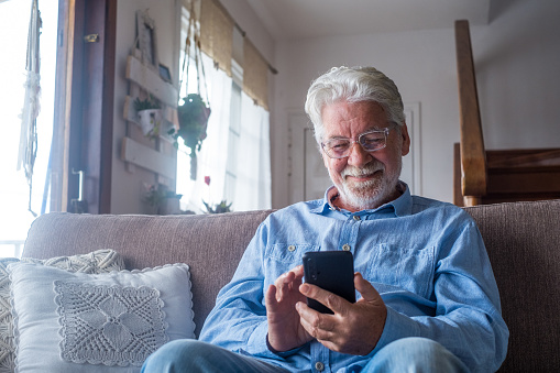 Old man smiling sitting on the sofa in the living room holding phone, enjoying using smartphone feeling satisfied sending messages, calling friends, surfing web online concept