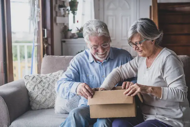 Photo of Happy mature aged older family couple unpacking carton box, satisfied with internet store purchase or unexpected gift, feeling excited of fast delivery shipping service, positive shopping experience.