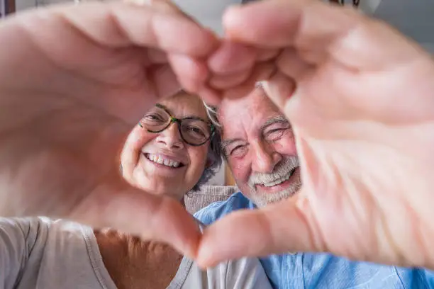 Photo of Close up portrait happy sincere middle aged elderly retired family couple making heart gesture with fingers, showing love or demonstrating sincere feelings together indoors, looking at camera.