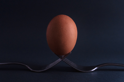 Chicken egg stands on two forks. Abstract still life, two forks and egg on a black background.
