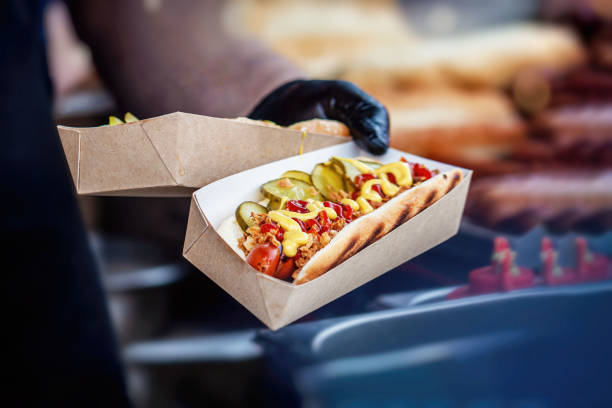 main du chef retenant la boîte en carton avec le hot-dog fraîchement préparé avec des additions savoureuses. barbecue, concept de gril - barbecue grill chef barbecue sausage photos et images de collection