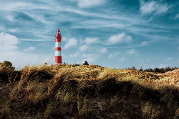 istock Tall red and white striped lighthouse on sand dunes in evening sunlight. Blue sky with white clouds. 1319868988