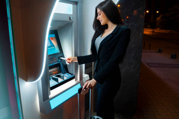 istock Caucasian woman with suitcase using ATM at night 1319876308