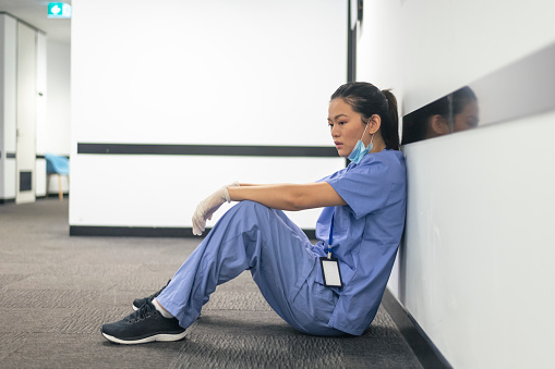 A female nurse of asian descent sits on the hospital hallway floor. The nurse appears exhausted and stressed after attending to patients with COVID-19 all day and night. She is emotionally drained and burnout from the long hours she has been working.