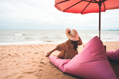 Young asian woman tourist solo travel tropical island. Relax on the beach cafe luxury pink concept in dress cloth and her arm hold a hat. Background with copy space.