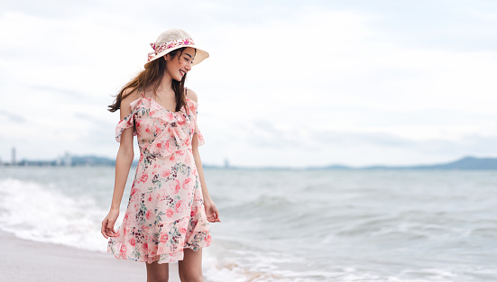 Young asian influencer smile woman walk and relax on the beach. Pattaya, Thailand. In pink flower dress and hat during summer season holiday. Background with copy space.