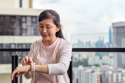 Middle aged Asian woman checking pulse on smart watch at balcony after exercising